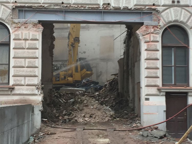 Foto: Sicht auf Baustelle im Innenhof eines Altbaugebäudes, Fassade mit Fenstern, Durchfahrt mit davorliegenden Schläuchen, im Hintergrund Abbruchkran auf einem Schutthaufen, rundherum Schutt, Ziegel und Staub 